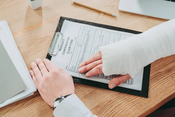 Cropped view of worker with broken arm in gypsum holding hands on compensation form in office, compensation concept — Stock Photo