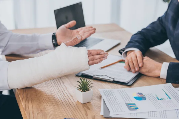 Vista recortada del trabajador con el brazo roto sentado en la mesa con documentos frente a hombre de negocios en la oficina, concepto de compensación - foto de stock