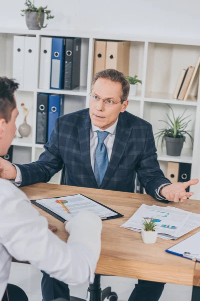 Homme d'affaires en veste bleue assis à table et vomissant les mains en face travailleur avec bras cassé dans le bureau, concept de compensation — Photo de stock