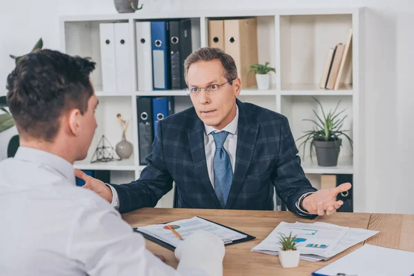 Businessman throwing up hands and siting at table opposite worker with broken arm in office, compensation concept — Stock Photo