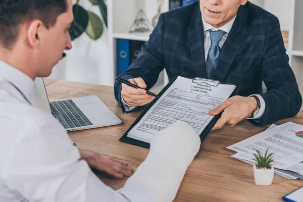 Businessman in blue jacket giving form for compensation claim to worker with broken arm at table in office, compensation concept — Stock Photo