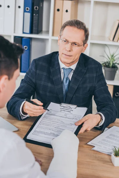 Hombre de negocios en chaqueta azul señalando en forma de reclamación de indemnización con pluma a trabajador con el brazo enyesado en la oficina, concepto de compensación - foto de stock