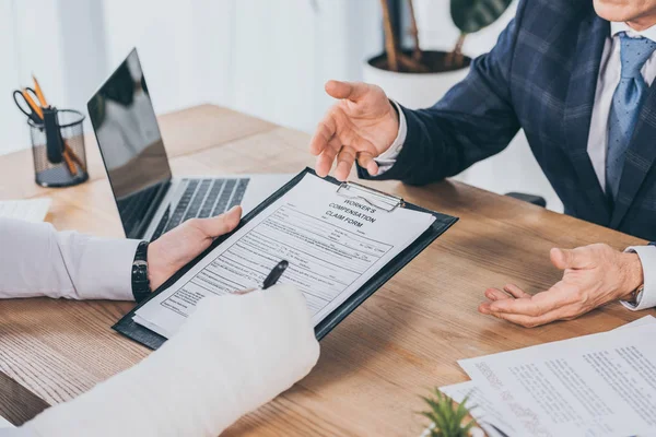 Cropped view of worker with broken arm signing form for compensation claim  opposite to businessman in blue jacket in office, compensation concept — Stock Photo