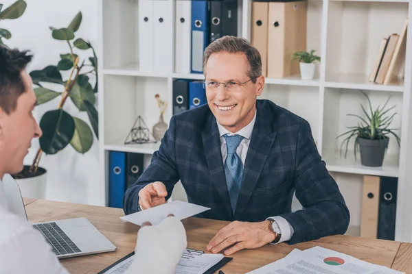 Worker with broken arm giving paper to businessman in blue jacket in office, compensation concept — Stock Photo