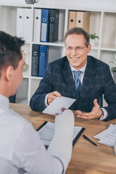 Businessman in blue jacket taking paper from worker with broken arm in office, compensation concept — Stock Photo