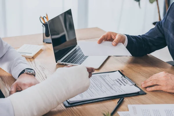 Vue recadrée d'un homme d'affaires assis à table et donnant une feuille de papier à un travailleur ayant un bras cassé au bureau, concept d'indemnisation — Photo de stock