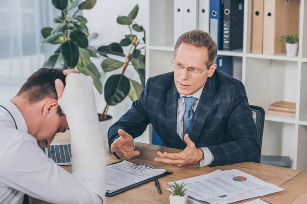 Worker with broken arm sitting at table and reading documents opposite to businessman in blue jacket in office, compensation concept — Stock Photo