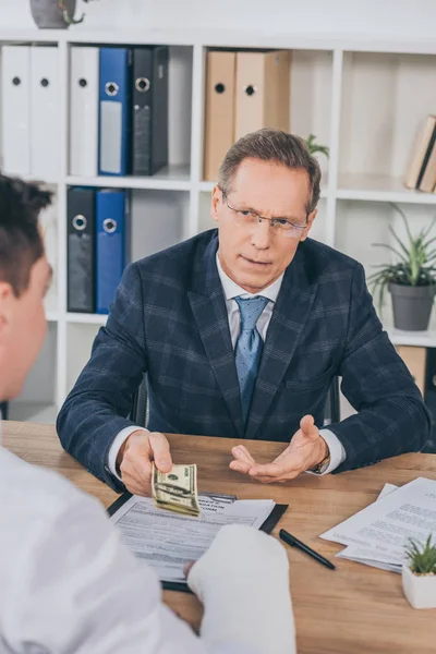 Businessman in blue jacket sitting at table and giving to worker with broken arm money in office, compensation concept — Stock Photo