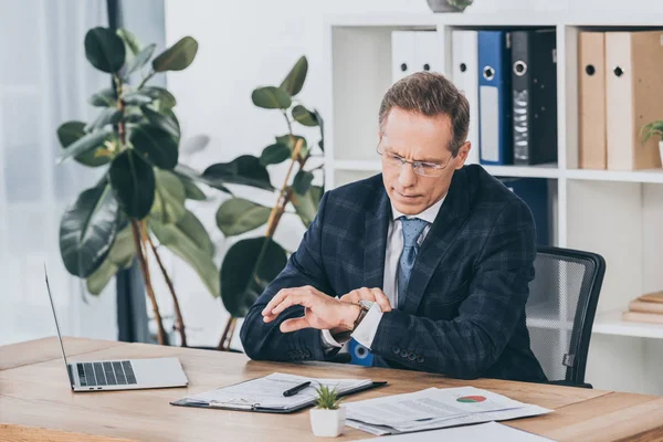Geschäftsmann in blauer Jacke sitzt mit Dokumenten am Tisch und schaut im Büro auf die Uhr, Vergütungskonzept — Stockfoto
