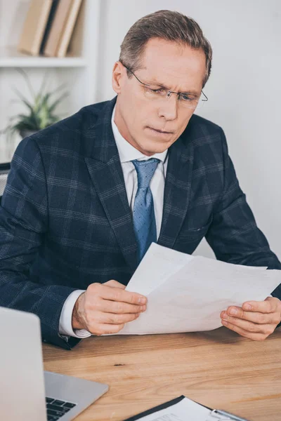 Uomo d'affari in giacca blu seduto a tavola con computer portatile e guardando attraverso il documento in ufficio, concetto di compensazione — Stock Photo