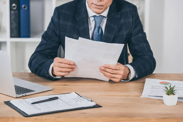 Vista cortada de empresário em casaco azul sentado à mesa e leitura de documentos no escritório, conceito de compensação — Fotografia de Stock