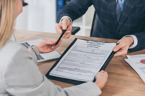 Vista recortada de hombre de negocios dando pluma mujer y forma para la reclamación de compensación sobre la mesa con el ordenador portátil en la oficina, concepto de compensación - foto de stock