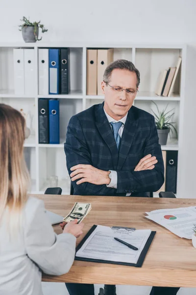 Middle aged businessman sitting at table with documents and looking on money giving by woman in office, compensation concept — Stock Photo