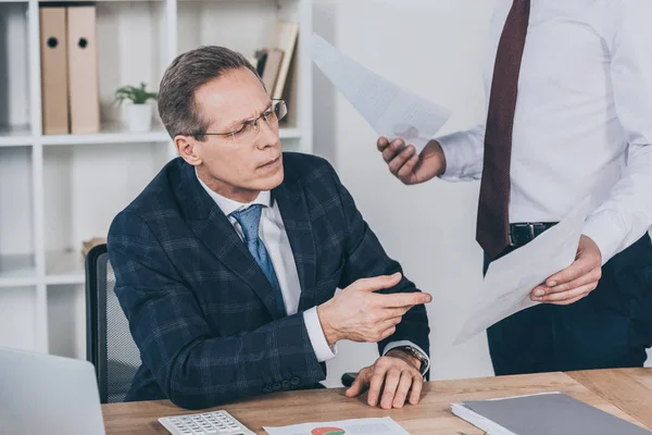 Middle aged businessman sitting at table wile worker standing and giving him documents in office, compensation concept — Stock Photo