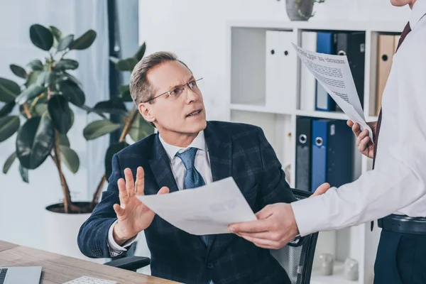 Middle aged businessman sitting at table, talking with worker standing near and holding documents in office, compensation concept — Stock Photo