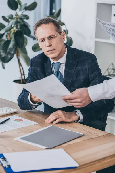Middle aged businessman sitting at table and readind document holding by worker in office, compensation concept — Stock Photo