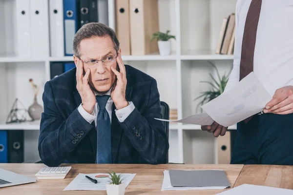 Середнього віку незадоволений бізнесмен, який сидить за столом працівник стоїть поруч з документами в офісі, концепція компенсації — стокове фото