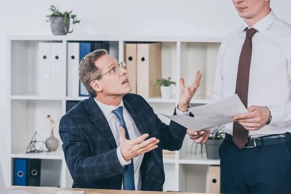 Middle aged dissatisfied businessman sitting at table and talking with worker standing near with documents in office, compensation concept — Stock Photo