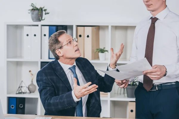 Hombre de negocios insatisfecho de mediana edad sentado en la mesa y hablando con el trabajador de pie cerca con documentos en la oficina, concepto de compensación - foto de stock