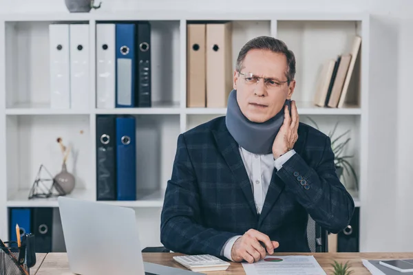 Hombre de negocios con corsé en el cuello sentado en la mesa, sintiendo dolor y sosteniendo el cuello en la oficina, concepto de compensación - foto de stock