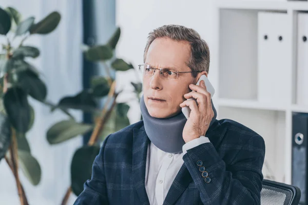 Бизнесмен в шейном корсете говорить на смартфоне в офисе, концепция компенсации — стоковое фото