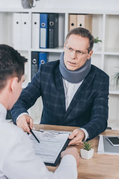 Businessman in neck brace sitting at table and pointing on form for compensation claim with finger to worker in office, compensation concept — Stock Photo