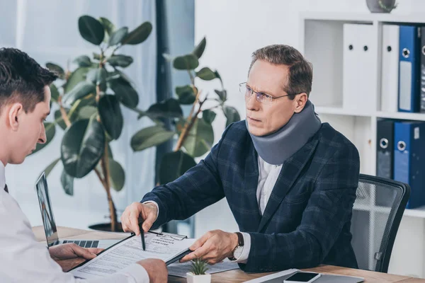 Businessman in neck brace sitting at table and pointing on form for compensation claim to worker in office, compensation concept — Stock Photo