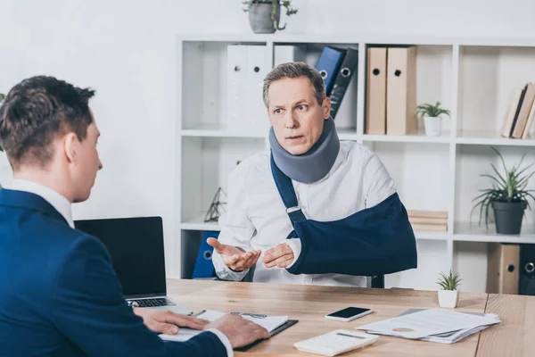 Trabajador de mediana edad en soporte para el cuello con el brazo roto sentado en la mesa y hablando con el hombre de negocios en chaqueta azul en la oficina, concepto de compensación - foto de stock