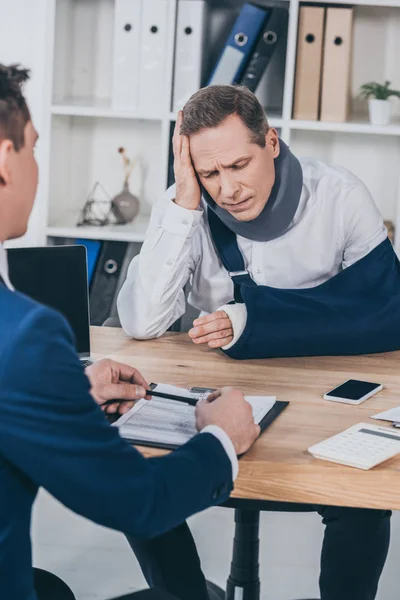 Upset worker in neck brace with broken arm sitting at table opposite businessman in blue jacket in office, compensation concept — Stock Photo