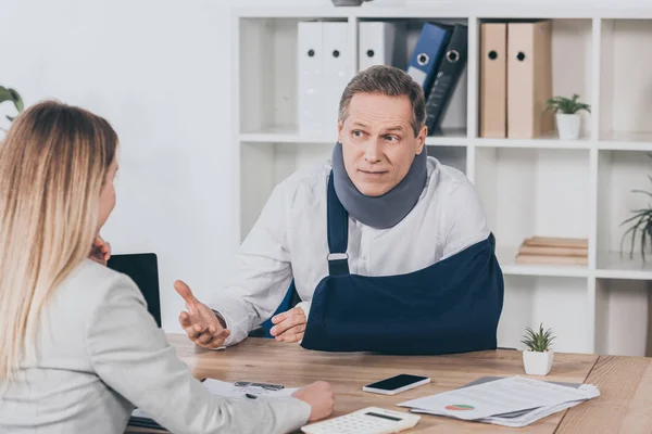 Middle aged worker in neck brace with broken arm sitting at table and talking to blond woman in office, compensation concept — Stock Photo