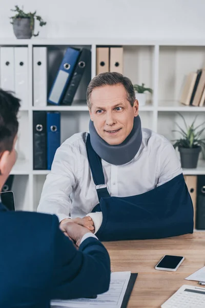 Geschäftsmann in blauer Jacke beim Händeschütteln mit Arbeiter in Nackenstütze und Armverband über Tisch im Büro, Vergütungskonzept — Stockfoto