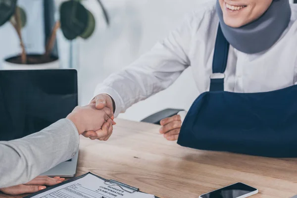 Vista ritagliata della donna che stringe la mano con lavoratore in tutore del collo e bendaggio del braccio sul tavolo in ufficio, concetto di compensazione — Foto stock