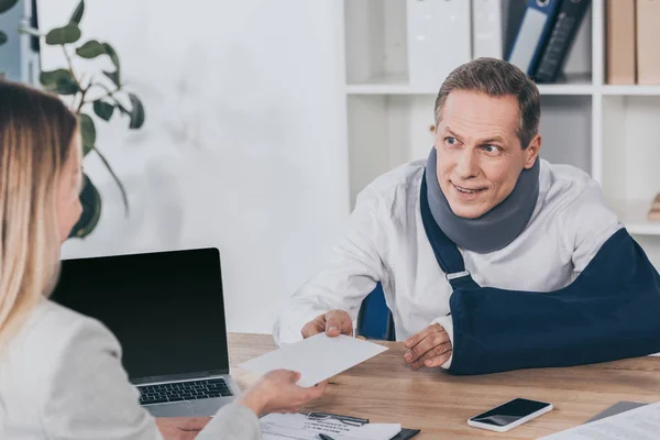 Worker in neck brace and arm bandage sitting at table and giving envelope to woman in office, compensation concept — Stock Photo