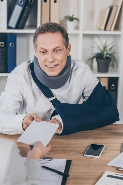 Worker in neck brace and arm bandage taking envelope by woman over table in office, compensation concept — Stock Photo