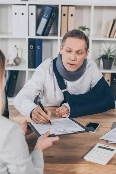 Travailleur dans le cou orthèse et bandage de bras signature tandis que la femme tenant tablette de papier sur la table au bureau, concept de rémunération — Photo de stock