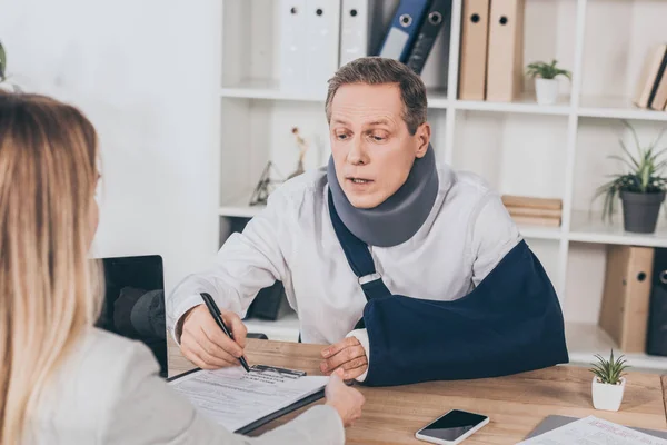 Worker in neck brace and arm bandage at table pointing with pen in document while woman sitting opposite in office, compensation concept — Stock Photo