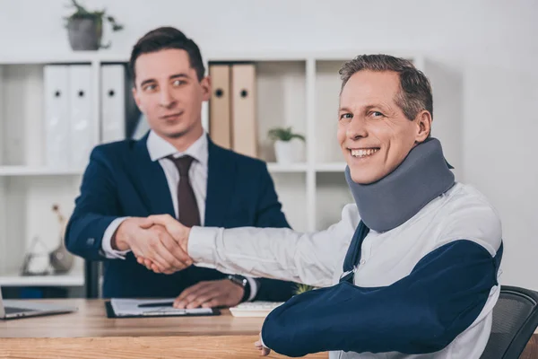 Hombre de negocios estrechando las manos con el trabajador en la abrazadera del cuello y el vendaje del brazo sobre la mesa en la oficina, concepto de compensación - foto de stock