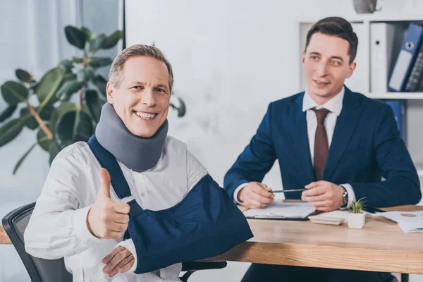 Hombre de negocios sentado en la mesa trabajador astuto en la abrazadera del cuello y el vendaje del brazo que muestra el pulgar hacia arriba en la oficina, concepto de compensación - foto de stock