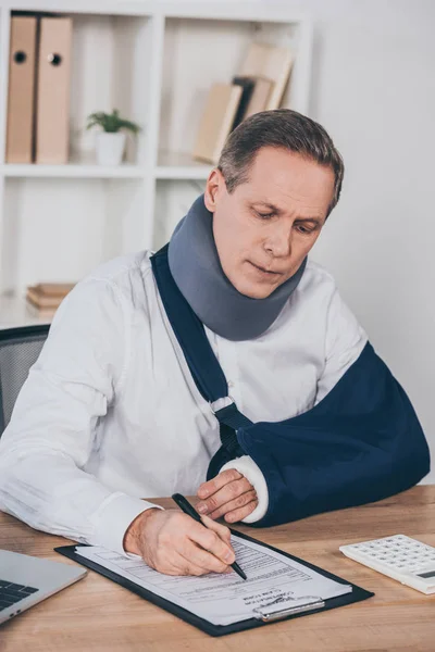Worker in neck brace and arm bandage sitting at table and writing with pen in office, compensation concept — Stock Photo