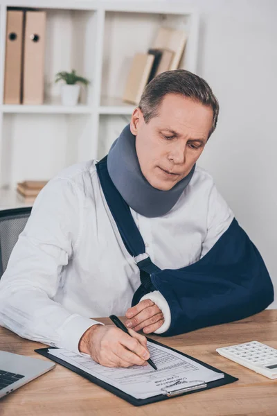 Trabajador en la abrazadera del cuello y el vendaje del brazo sentado en la mesa y la escritura con la pluma en la oficina, concepto de compensación - foto de stock