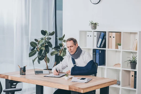 Worker in neck brace and arm bandage sitting at table and carefully writing in office, compensation concept — Stock Photo