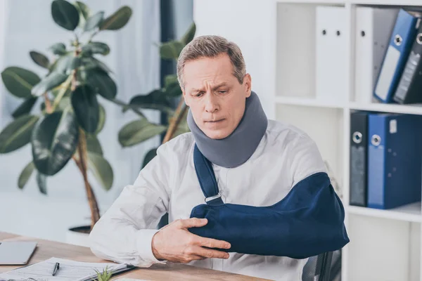 Middle aged worker holding broken arm wile sitting at table with document in office, compensation concept — Stock Photo