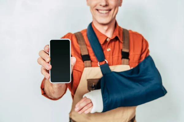 Vista ritagliata del riparatore con fasciatura del braccio che mostra smartphone vuoto con schermo vuoto su sfondo bianco — Foto stock