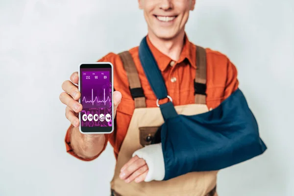 Обрезанный вид ремонтника со сломанной рукой и бинтом, показывающий смартфон на белом фоне — стоковое фото