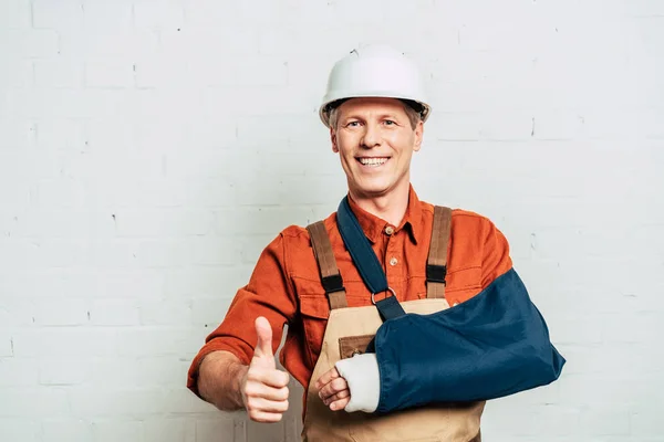 Reparador con el brazo vendaje de pie y mostrando pulgar hacia arriba sobre fondo texturizado blanco - foto de stock