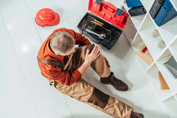 Top view of repairman sitting on floor and holding injured knee surrounding by equipment in office — Stock Photo