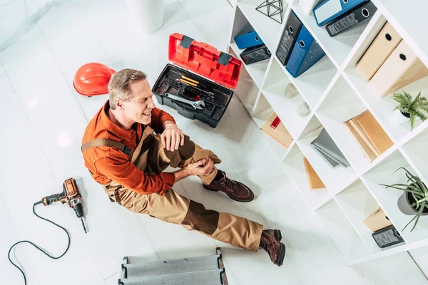 Ansicht von oben: Reparateur sitzt mit lädiertem Knie im Büro auf dem Boden — Stockfoto