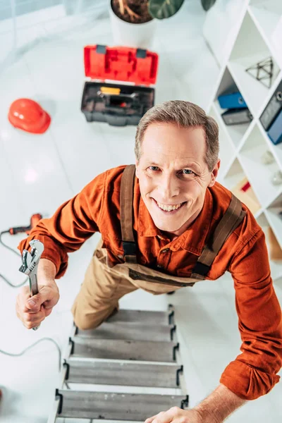 Vista superior del reparador de escalada en escalera con llave inglesa y sonriendo en la oficina - foto de stock