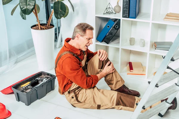 Repairman sitting on floor and holding injured knee surrounding by equipment in office — Stock Photo