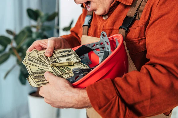 Vista ritagliata di riparatore sorpreso che tiene hardhat con i soldi in ufficio — Foto stock